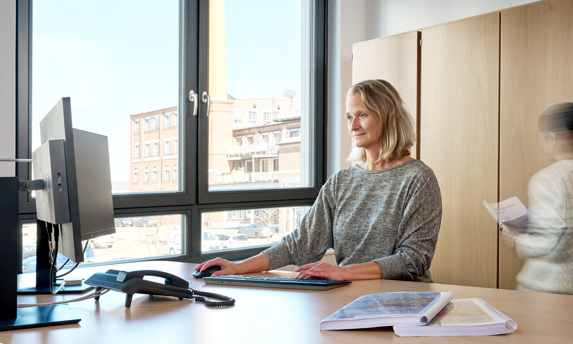 Lächelnde Frau am Schreibtisch vor Fensterfront bei der Arbeit